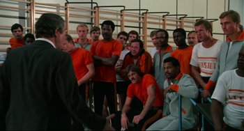 Movie still from “Rollerball” (1975), directed by Norman Jewison – A group of men standing next to each other in orange shirts; Medium shot, Over the shoulder angle