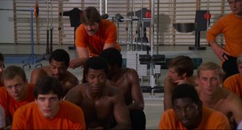 Movie still from “Rollerball” (1975), directed by Norman Jewison – A group of men sitting in a gym with a trainer looking on; Medium shot, Over the shoulder angle
