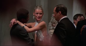 Movie still from “Rollerball” (1975), directed by Norman Jewison – A woman in a silver dress getting ready for an event with two other men; Medium shot, Over the shoulder angle