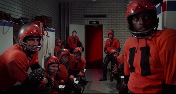 Movie still from “Rollerball” (1975), directed by Norman Jewison – A group of men in red football uniforms in a room; Medium shot, High angle