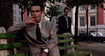 Movie still from “Romeo Is Bleeding” (1993), directed by Peter Medak – A man smoking a cigarette on a park bench with another man standing behind him; Medium shot, Low angle