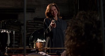Movie still from “Romeo Is Bleeding” (1993), directed by Peter Medak – A woman holding a gun in a room with other people; Medium shot, Over the shoulder angle