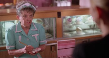 Movie still from “Romy and Michele's High School Reunion” (1997), directed by David Mirkin – An older woman in a blue dress holding a white object; Medium shot, Over the shoulder angle