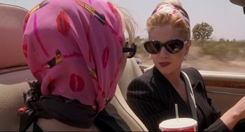 Movie still from “Romy and Michele's High School Reunion” (1997), directed by David Mirkin – A woman in sunglasses and a pink scarf is sitting in a car with another woman; Close Up shot, Over the shoulder angle