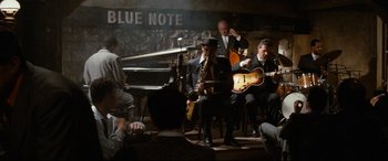 Movie still from “'Round Midnight” (1986), directed by Bertrand Tavernier – A group of people sitting around a room playing musical instruments; Wide shot, High angle