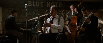 Movie still from “'Round Midnight” (1986), directed by Bertrand Tavernier – A man sitting down playing a saxophone in front of another man; Wide shot, High angle
