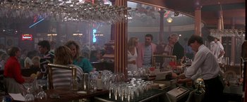 Movie still from “Roxanne” (1987), directed by Fred Schepisi – A group of people standing around a bar with glasses; Wide shot, Over the shoulder angle