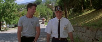 Movie still from “Roxanne” (1987), directed by Fred Schepisi – Two men walking down a street with trees in the background; Medium shot, Low angle