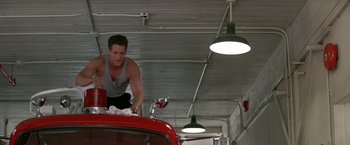 Movie still from “Roxanne” (1987), directed by Fred Schepisi – A man in grey shirt and black shorts working on a red car; Medium shot, Low angle