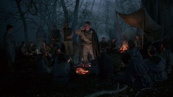 Movie still from “Royal Deceit” (1994), directed by Gabriel Axel – A group of people sitting around a fire in the woods; Wide shot, High angle