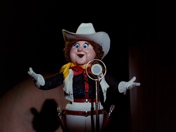 Movie still from “Rudolph and Frosty's Christmas in July” (1979), directed by Jules Bass – A doll is wearing a cowboy outfit while holding a microphone; Medium shot, Low angle