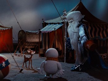 Movie still from “Rudolph and Frosty's Christmas in July” (1979), directed by Jules Bass – A scene of a scene where a person is standing in front of an animal and another animal is standing in front of a tent; Wide shot, High angle