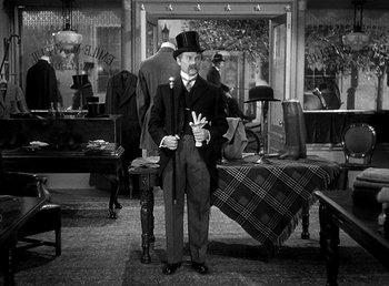 Movie still from “Ruggles of Red Gap” (1935), directed by Leo McCarey – A man in a top hat and suit standing in front of a table; Wide shot, High angle