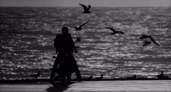 Movie still from “Rumble Fish” (1983), directed by Francis Ford Coppola – A person sitting on a motor scooter near the ocean; Wide shot, Low angle