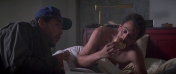 Movie still from “Running Scared” (1986), directed by Peter Hyams – A man eating a hot dog in bed while another man watches; Medium shot, High angle