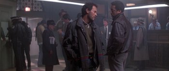 Movie still from “Running Scared” (1986), directed by Peter Hyams – Two men are standing in a group of people; Medium shot, Over the shoulder angle