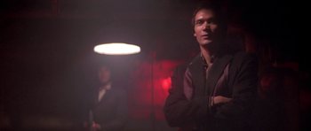 Movie still from “Running Scared” (1986), directed by Peter Hyams – A man standing next to another man in a dark room; Medium shot, Low angle