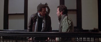 Movie still from “Running Scared” (1986), directed by Peter Hyams – Two men talking to each other while standing next to a railing; Medium shot, Over the shoulder angle