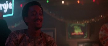 Movie still from “Running Scared” (1986), directed by Peter Hyams – A man smiling at the camera in front of a television; Close Up shot, Low angle