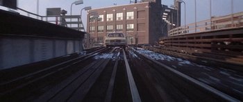 Movie still from “Running Scared” (1986), directed by Peter Hyams – A car driving down a train track next to a building; Extreme Wide shot, High angle