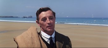 Movie still from “Ryan's Daughter” (1970), directed by David Lean – A man wearing a suit and tie standing on a beach; Close Up shot, Over the shoulder angle