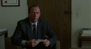 Movie still from “Safe” (1995), directed by Todd Haynes – A man in a suit sitting in a chair holding a business card; Close Up shot, Over the shoulder angle