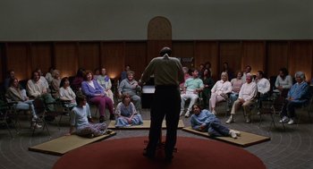 Movie still from “Safe” (1995), directed by Todd Haynes – A group of people sitting in a circle with a man standing in front of them; Wide shot, High angle