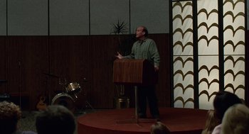 Movie still from “Safe” (1995), directed by Todd Haynes – A man standing at a podium on a stage; Medium shot, Low angle