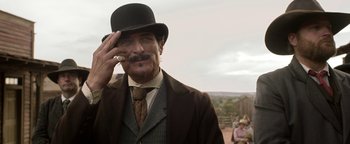 Movie still from “Godless” (2017), directed by Scott Frank – A man in a top hat smoking a cigarette while wearing a suit; Medium shot, Low angle