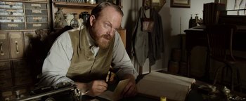 Movie still from “Godless” (2017), directed by Scott Frank – A man sitting at a table writing on a piece of paper; Medium shot, Over the shoulder angle