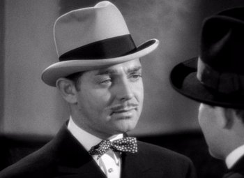 Movie still from “San Francisco” (1936), directed by W.S. Van Dyke – A black and white photo of a man wearing a hat and a bow tie; Close Up shot, Over the shoulder angle