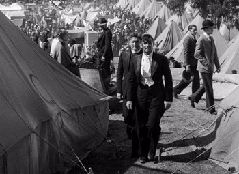 Movie still from “San Francisco” (1936), directed by W.S. Van Dyke – A man and a woman are walking through a tent city; Wide shot, High angle