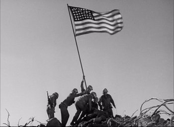 Movie still from “Sands of Iwo Jima” (1949), directed by Allan Dwan – A group of men standing on top of a hill holding an american flag; Wide shot, Low angle