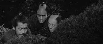 Movie still from “Sanjuro” (1962), directed by Akira Kurosawa – Two people with hair on their heads are sitting in a bush; Medium shot, High angle