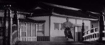 Movie still from “Sanjuro” (1962), directed by Akira Kurosawa – A man standing in front of a building holding a sword; Wide shot, Low angle