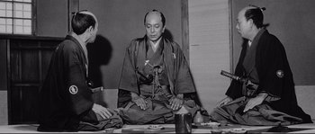Movie still from “Sanjuro” (1962), directed by Akira Kurosawa – A man sitting on the ground in front of two other men; Medium shot, Low angle
