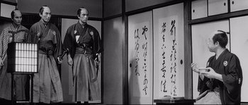 Movie still from “Sanjuro” (1962), directed by Akira Kurosawa – A man standing in front of a wall with japanese writing on it; Medium shot, Low angle