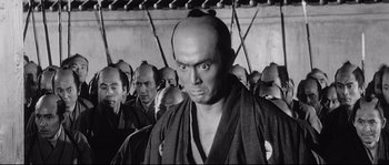 Movie still from “Sanjuro” (1962), directed by Akira Kurosawa – A man with a bald head in front of a group of men; Medium shot, Low angle
