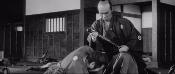 Movie still from “Sanjuro” (1962), directed by Akira Kurosawa – A man is holding a sword while another man is kneeling over him; Medium shot, Over the shoulder angle