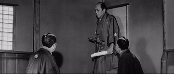 Movie still from “Sanjuro” (1962), directed by Akira Kurosawa – An old photo of a man holding a sword in front of two other men; Medium shot, Low angle