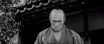 Movie still from “Sanjuro” (1962), directed by Akira Kurosawa – A man with a shaved head wearing a kimono; Close Up shot, Low angle