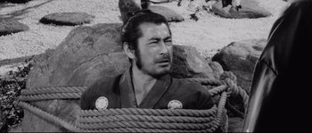 Movie still from “Sanjuro” (1962), directed by Akira Kurosawa – A man with a beard is sitting on a rope; Close Up shot, High angle