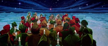 Movie still from “Santa Claus” (1985), directed by Jeannot Szwarc – A group of people dressed as elves are gathered around candles; Wide shot, High angle