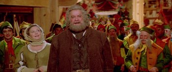 Movie still from “Santa Claus” (1985), directed by Jeannot Szwarc – A man with a long beard standing in front of a group of people; Medium shot, Low angle
