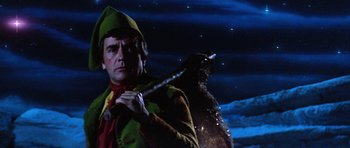 Movie still from “Santa Claus” (1985), directed by Jeannot Szwarc – A man dressed as a christmas elf holding a stick; Medium shot, Low angle
