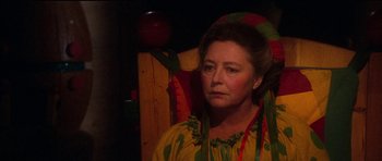 Movie still from “Santa Claus” (1985), directed by Jeannot Szwarc – A woman in a yellow and green dress sitting in a chair; Close Up shot, Over the shoulder angle
