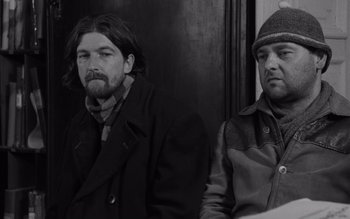 Movie still from “Satantango” (1994), directed by Béla Tarr – Two men sitting next to each other on a train; Medium shot, Low angle