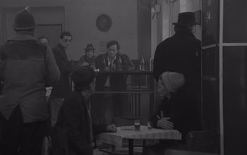 Movie still from “Satantango” (1994), directed by Béla Tarr – A black and white photo of a group of people sitting at a table; Wide shot, High angle