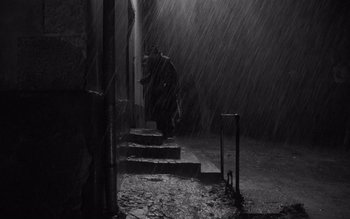 Movie still from “Satantango” (1994), directed by Béla Tarr – A man standing on some steps in the rain; Wide shot, Low angle