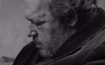 Movie still from “Satantango” (1994), directed by Béla Tarr – An older man with a goatee is looking down at his face; Close Up shot, Low angle